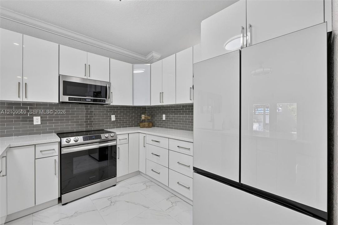 Kitchen featuring high-gloss white cabinetry, light-toned stone-finish countertops, grey subway tile backsplash, stainless steel appliances, and large-format white tile flooring with grey veining