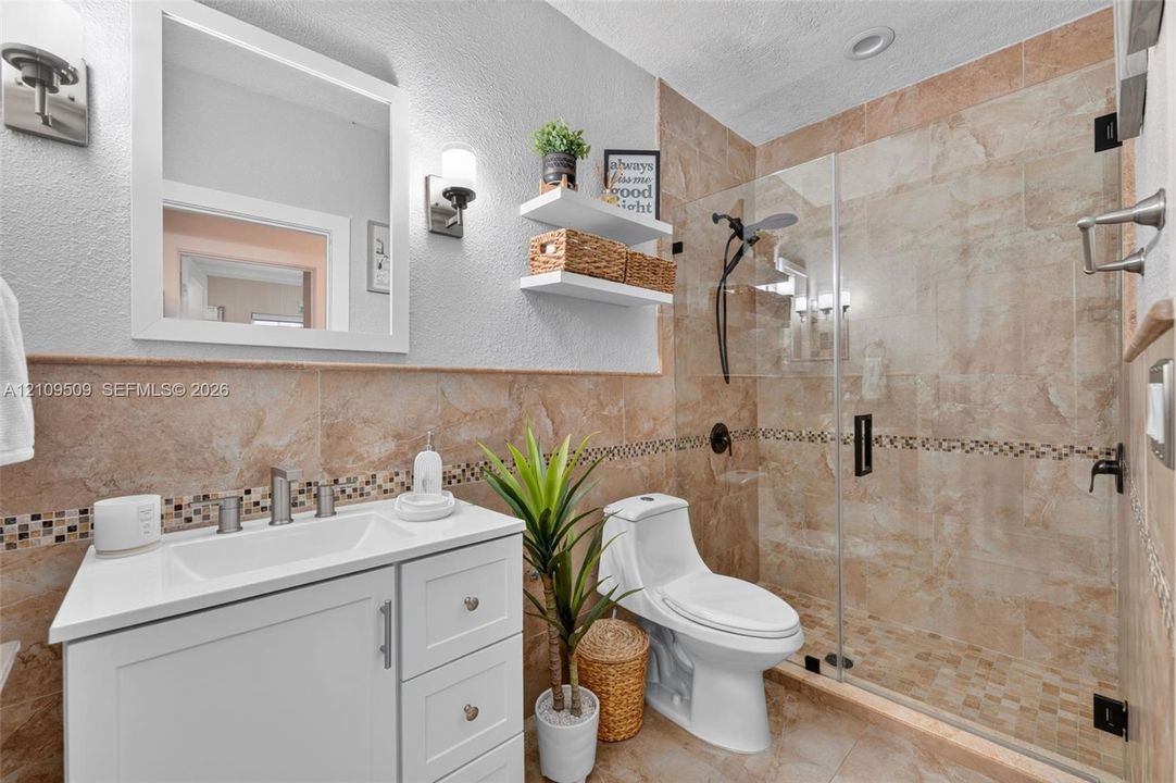Bathroom featuring a white vanity with an integrated sink, a glass-enclosed walk-in shower with mosaic tile accents, wall-mounted lighting, and neutral stone-finish wall and floor tiling