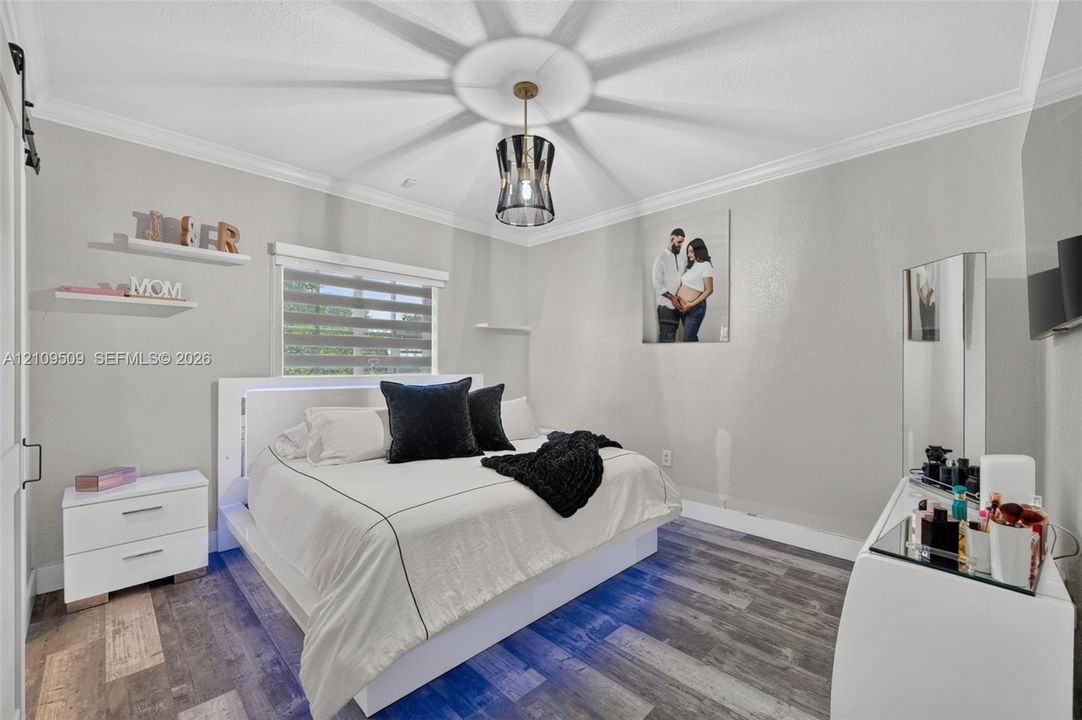 Bedroom featuring wood-finish flooring, crown molding, tiered window shades, and a ceiling-mounted pendant light fixture