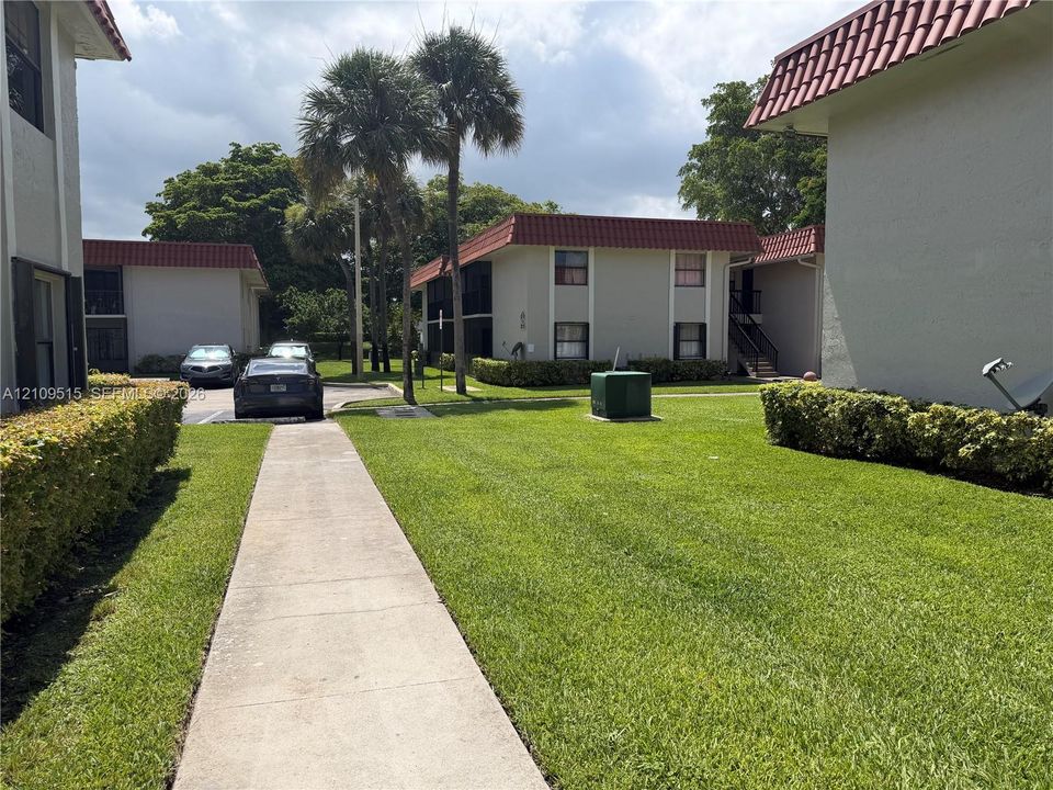 Exterior grounds featuring a concrete walkway, manicured lawn, perimeter hedges, and palm trees with multi-story buildings topped with red tile roofing