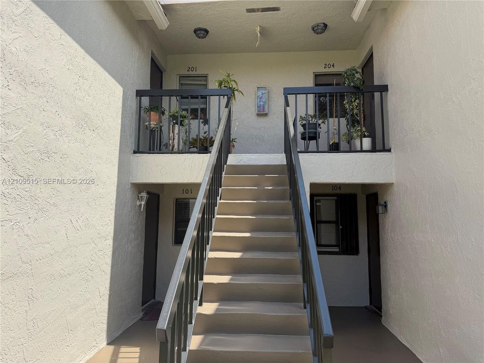 Exterior stairwell featuring stucco-finished walls, metal railings, and dual upper-level balconies with exterior lighting fixtures