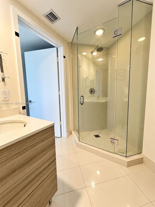 Glass-enclosed corner shower with a polished metal showerhead, vanity with wood-finish cabinetry and light-colored countertop, and large-format tile flooring