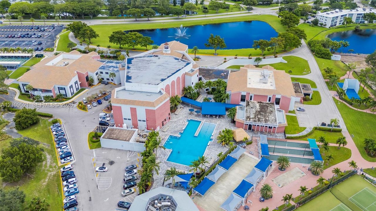 Aerial perspective featuring a multi-building property complex with a large swimming pool, paved surface parking areas, multi-court recreational facilities, and manicured grounds centered around a water feature with a fountain