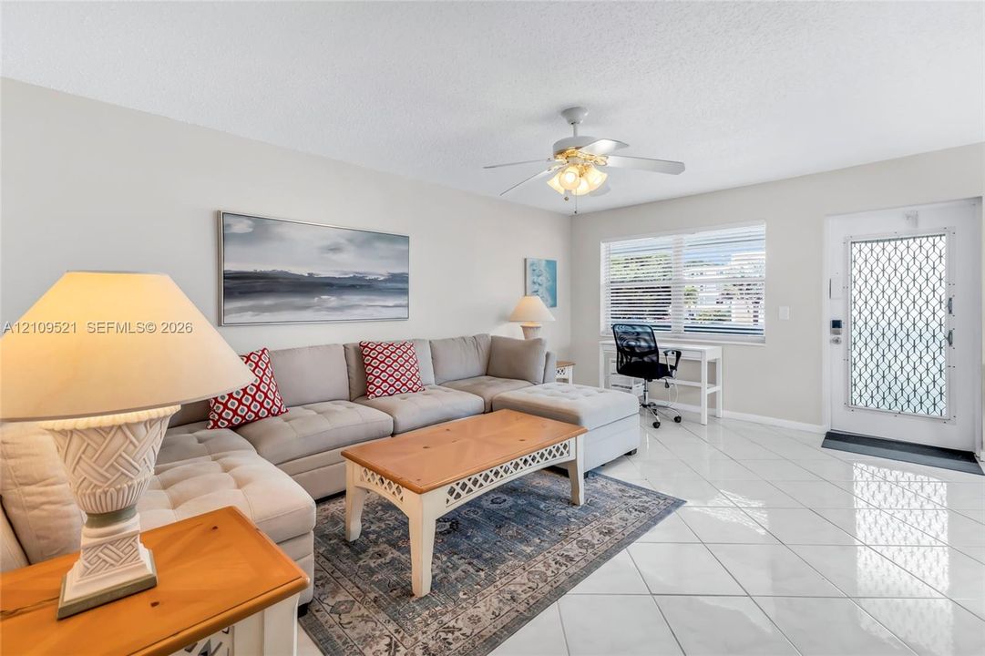 Living area featuring large-format ceramic tile flooring, a multi-blade ceiling fan with integrated lighting, and a window with horizontal blinds