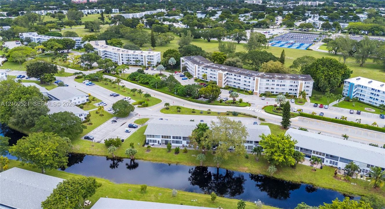 Aerial perspective of low-rise residential structures featuring exterior balconies, surface parking, mature landscaping, and waterfront grounds