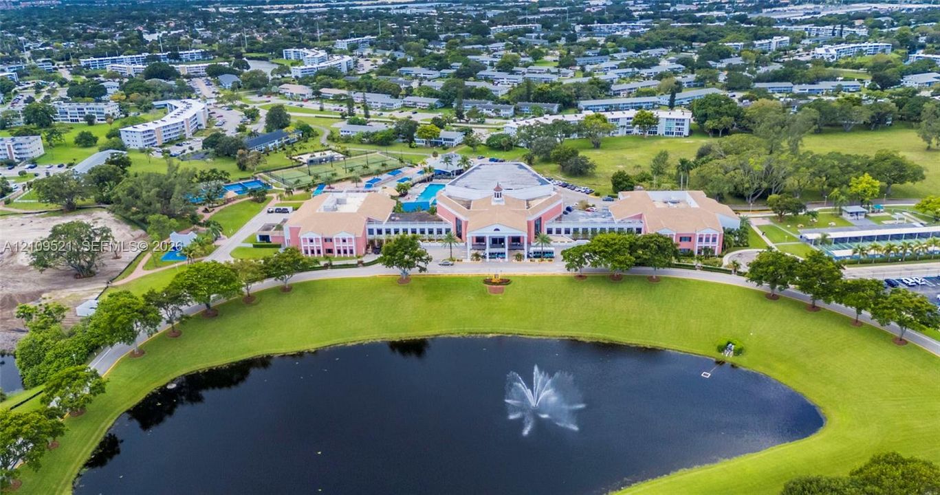 Aerial perspective of a multi-story building featuring a central cupola, expansive manicured grounds, a decorative fountain, and a large body of water