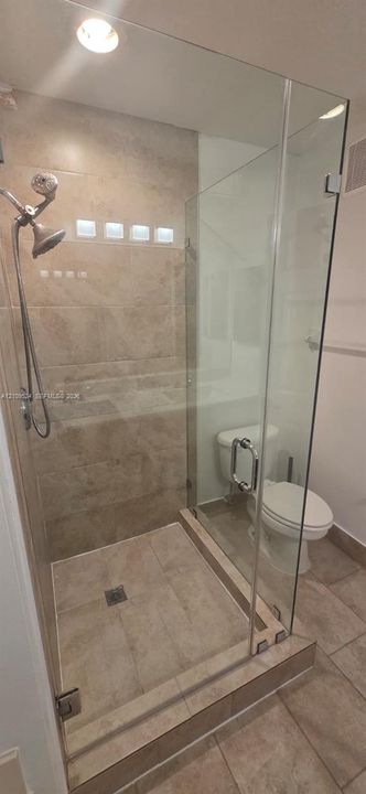 Walk-in shower featuring a frameless glass enclosure, neutral tile surround, handheld showerhead, and a tiled curb
