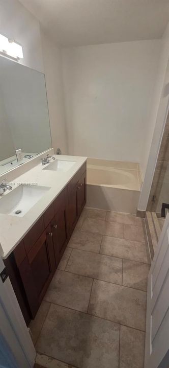 Double vanity featuring wood-finish cabinetry, solid surface countertops, dual undermount sinks, and a large-format wall mirror with a tiled floor and built-in soaking tub