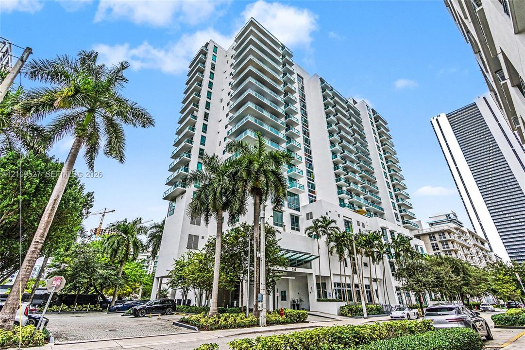 High-rise residential building featuring white concrete facade, glass-railed balconies, and ground-level landscaping with mature palm trees
