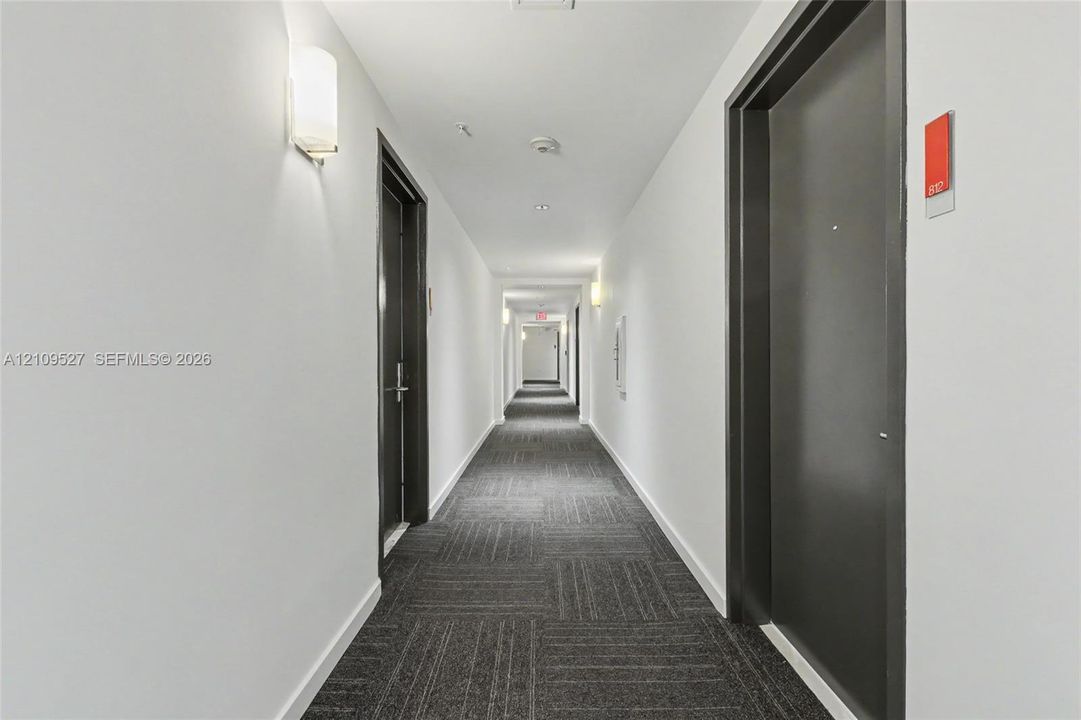Interior hallway featuring dark-finished doors, wall-mounted sconce lighting, and geometric-patterned carpet tile flooring