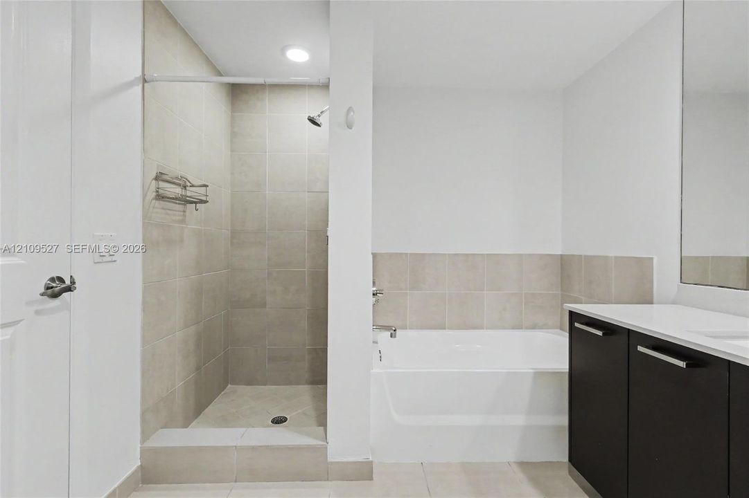Walk-in shower with neutral tile surround, deep soaking tub, dark wood-finish cabinetry, and solid surface countertop