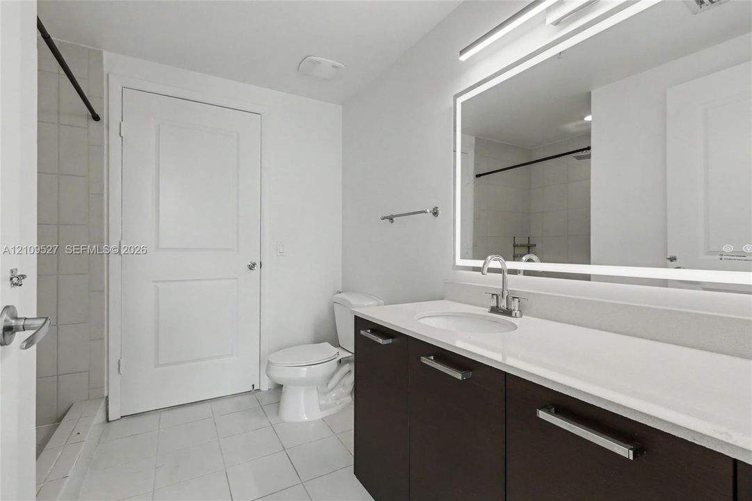 Bathroom featuring a dark wood-finish vanity with a light-toned solid surface countertop, an integrated oval sink, a large LED-backlit mirror, and tile flooring