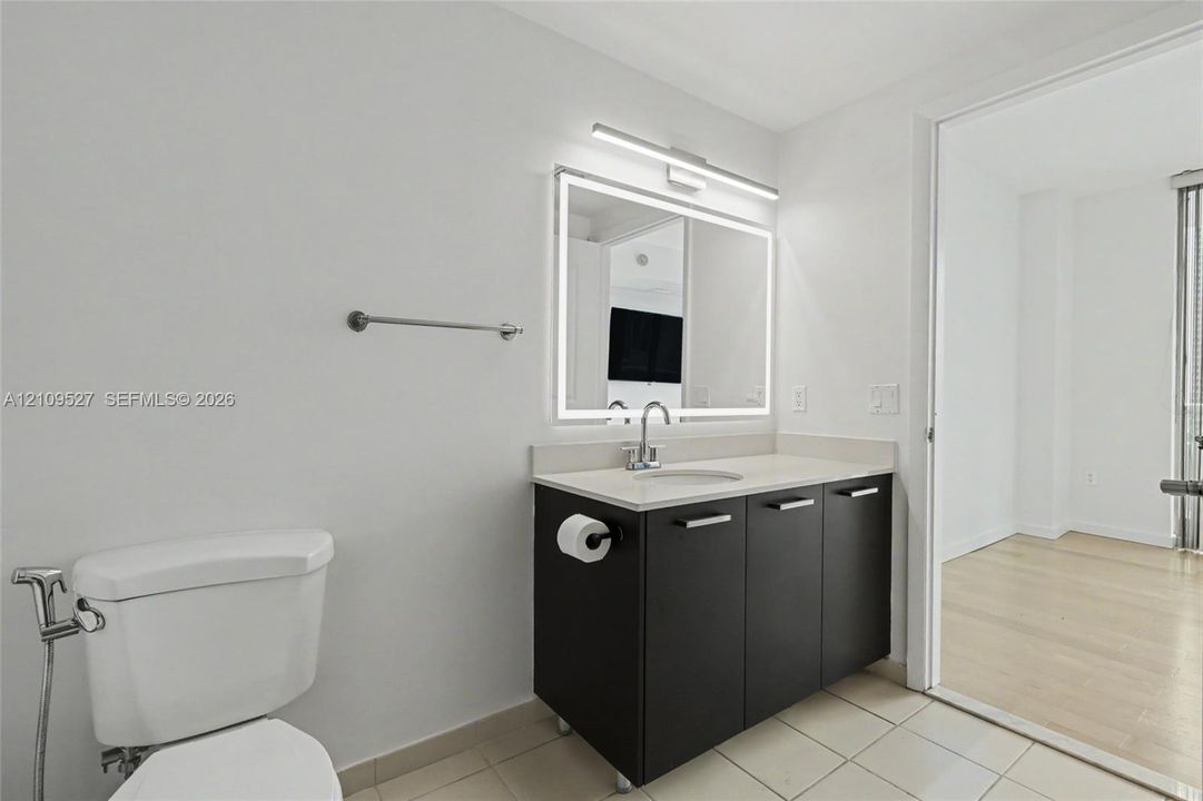 Dark-toned vanity with a light-toned solid surface countertop, integrated sink, wall-mounted LED-backlit mirror, brushed metal hardware, and ceramic tile flooring