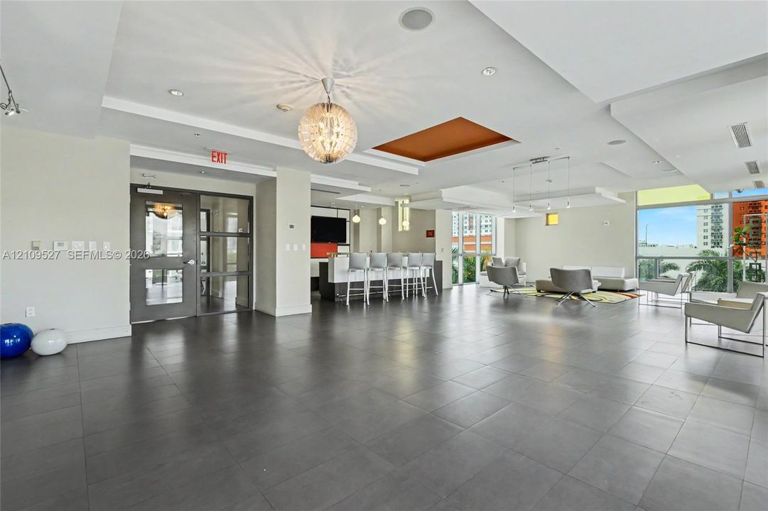 Large open-concept space featuring large-format tile flooring, tray ceilings with recessed lighting, a modern wet bar with built-in cabinetry, and expansive windows