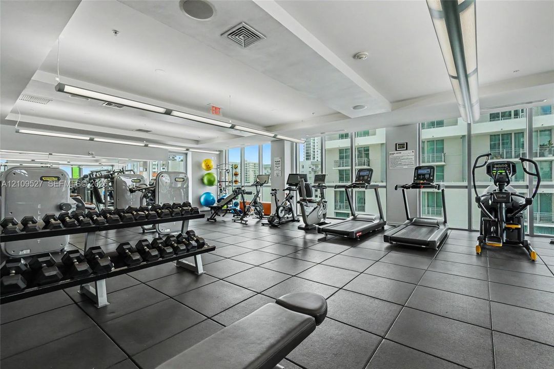 Fitness center featuring floor-to-ceiling windows, rubberized modular tile flooring, recessed architectural lighting, and mirrored walls