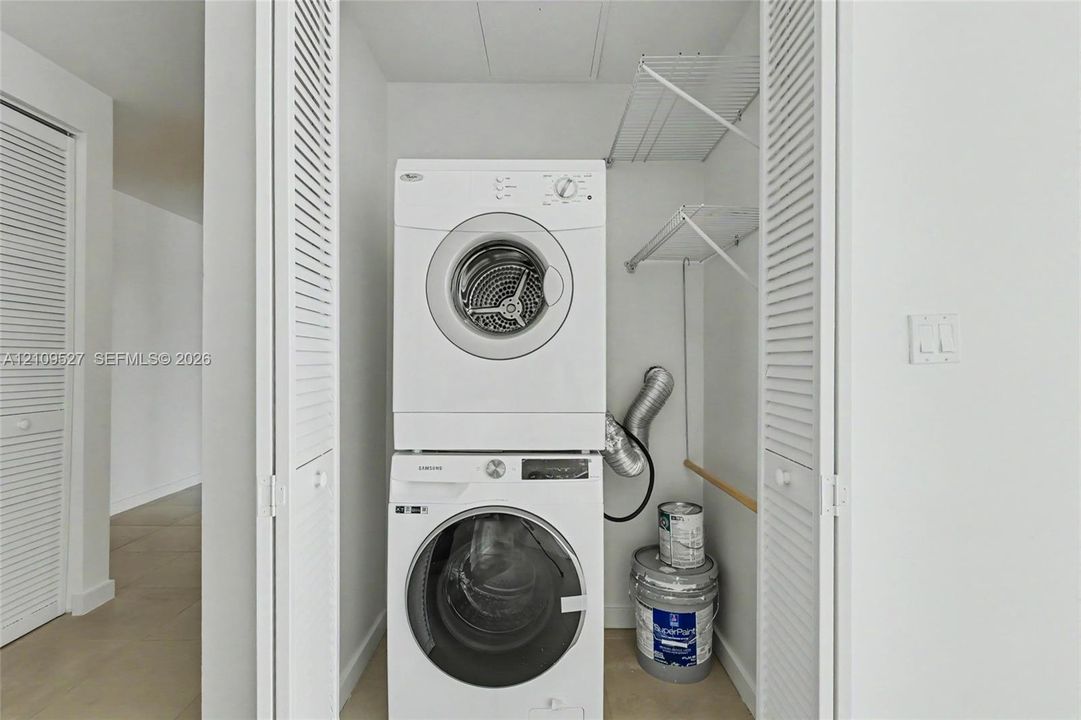 Laundry closet featuring stacked front-load laundry appliances and white louvered bifold doors with built-in wire shelving