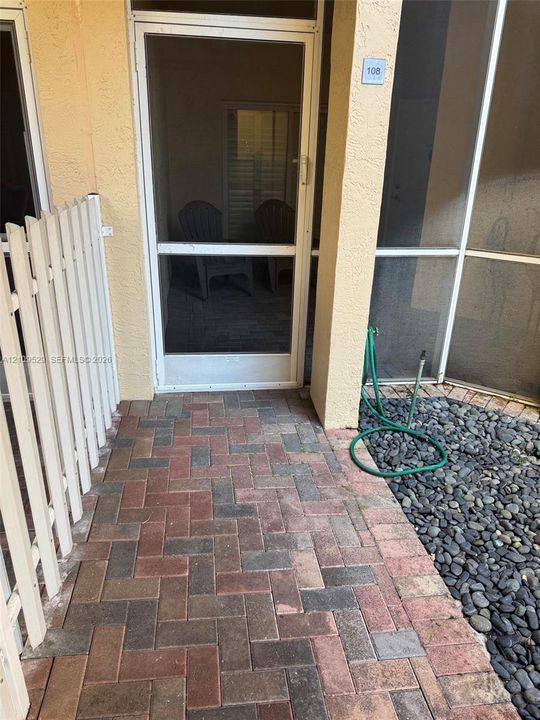 Entrance featuring a herringbone-patterned paver walkway, white vertical picket fencing, a screen door, and decorative stone ground cover