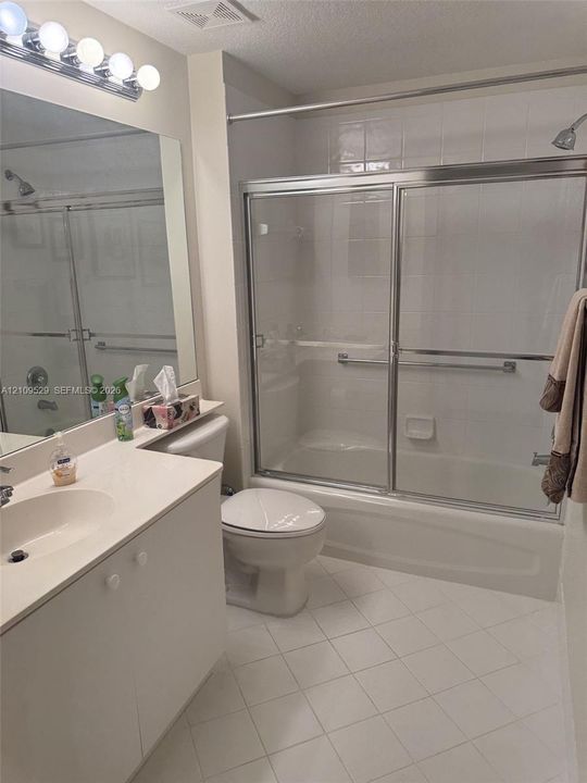 Full-width vanity with an integrated sink, polished chrome light fixture, large wall-mounted mirror, white tile flooring, and a shower-tub combination featuring sliding glass doors