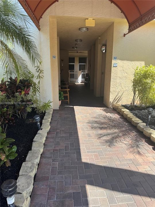Covered entryway featuring a herringbone-patterned paver walkway, decorative scalloped awning, tiered stone edging, and integrated landscape lighting
