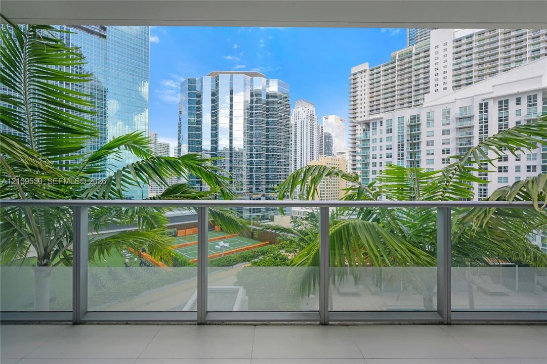 Covered balcony featuring tile flooring and a glass and metal railing, overlooking an outdoor tennis court with manicured palm trees