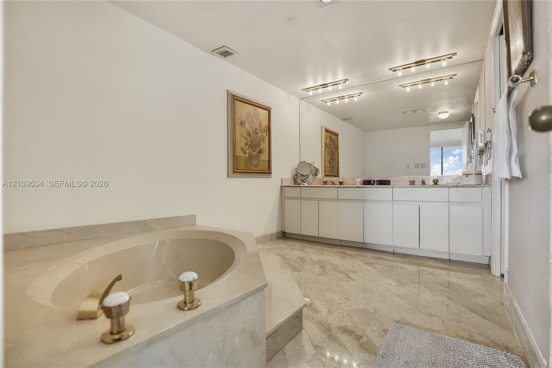 Bathroom featuring stone tile flooring, built-in vanity with white cabinetry, stone-surround soaking tub with brass-tone fixtures, and expansive wall-to-wall mirror