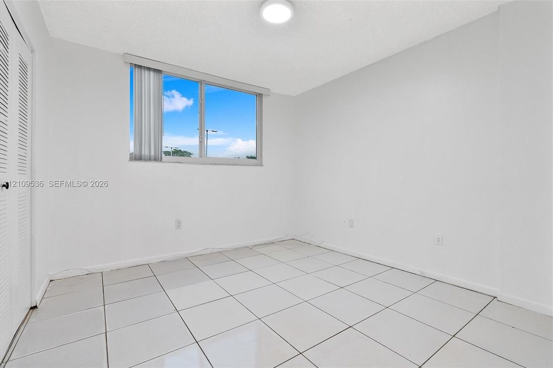 Room featuring large-format ceramic tile flooring, neutral walls, flush-mount ceiling lighting, and a window with vertical blinds, finished with louvered closet doors