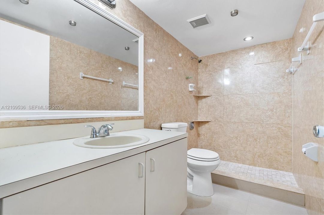 Bathroom featuring floor-to-ceiling stone-finish wall tile, an expansive vanity with a solid-surface countertop, an integrated sink, a large wall-mounted mirror, and a walk-in shower with built-in corner shelving