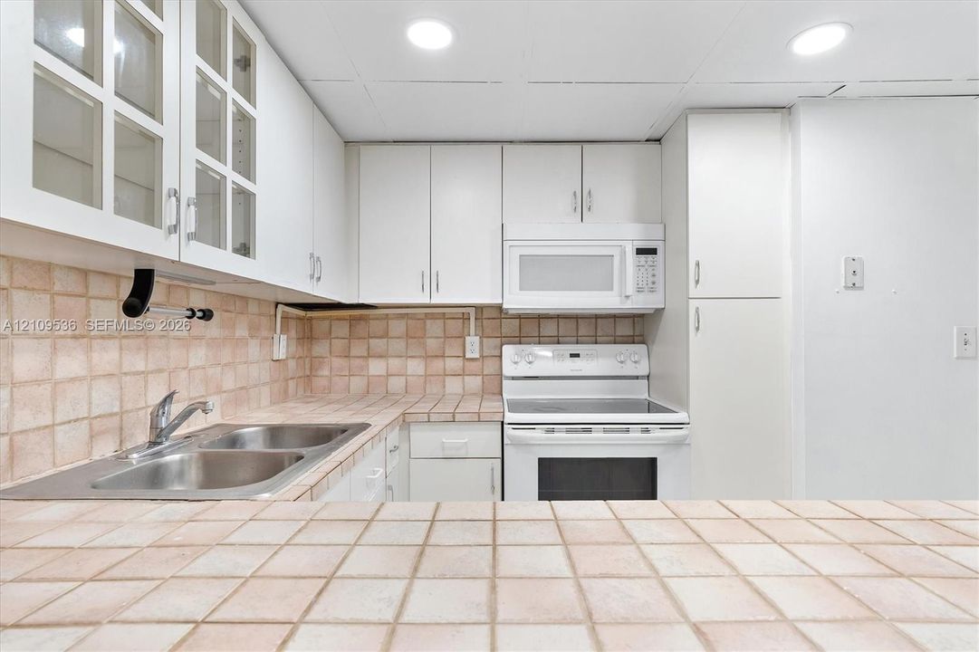 Kitchen featuring white cabinetry with glass-panel upper doors, tiled countertops and backsplash, dual-basin stainless steel sink, and white appliances including a built-in microwave and range