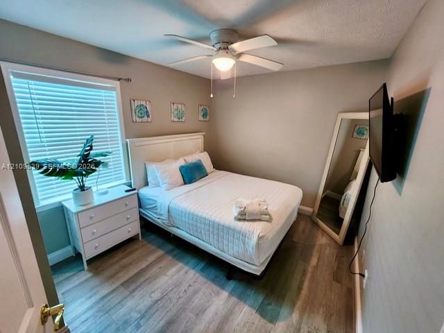 Wood-finish flooring, ceiling fan with integrated lighting, window with blinds, wall-mounted television, and a white bed frame