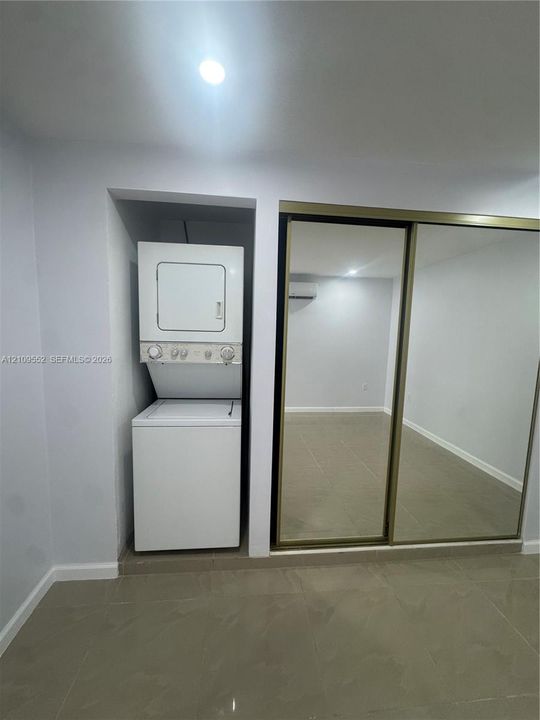 Recessed laundry alcove featuring stacked washer and dryer, floor-to-ceiling mirror-panel closet doors with metal trim, tile flooring, and wall-mounted ductless climate control unit