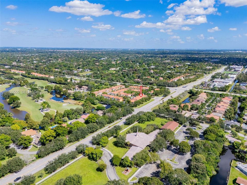 Aerial perspective of residential community featuring multi-story structures with tile roofing, surrounded by mature trees, water features, and expansive green space