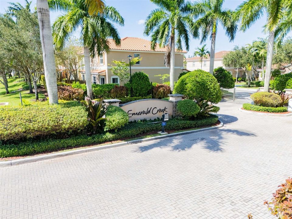 Community entrance featuring a stone monument sign, paver-stone roadway, manicured landscaping, and mature palm trees