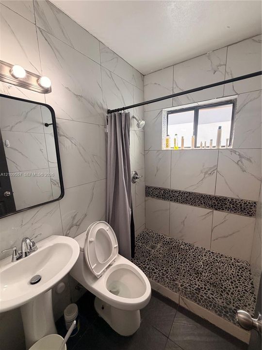 Bathroom featuring large-format veined wall tile, a pedestal sink, a walk-in shower with stone pebble flooring, a decorative stone tile inlay, and a rectangular vanity mirror