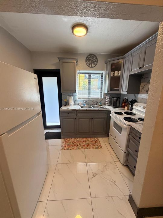 Kitchen featuring gray cabinetry, large-format tile flooring, stone-finish backsplash, dual-basin stainless steel sink, and glass-front upper cabinet