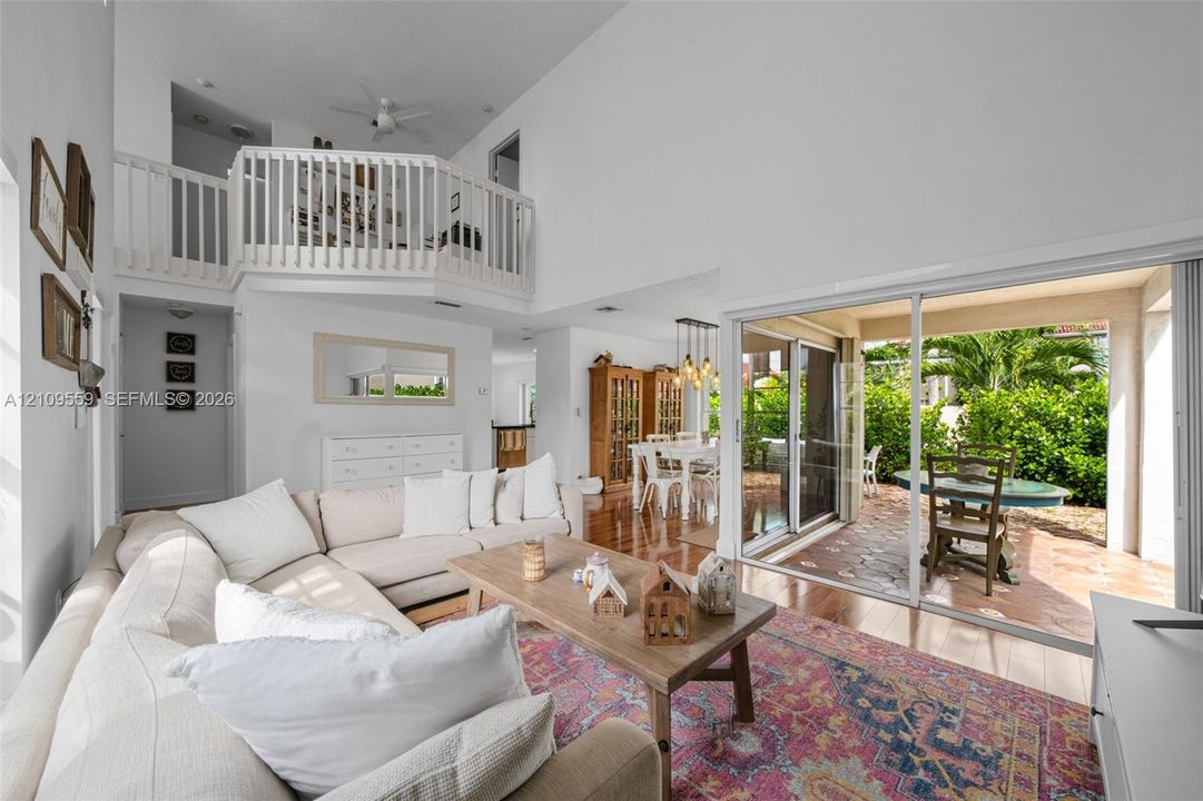 Living area with vaulted ceilings, elevated mezzanine loft with white railing, wood-finish flooring, and large sliding glass doors opening to a tiled patio