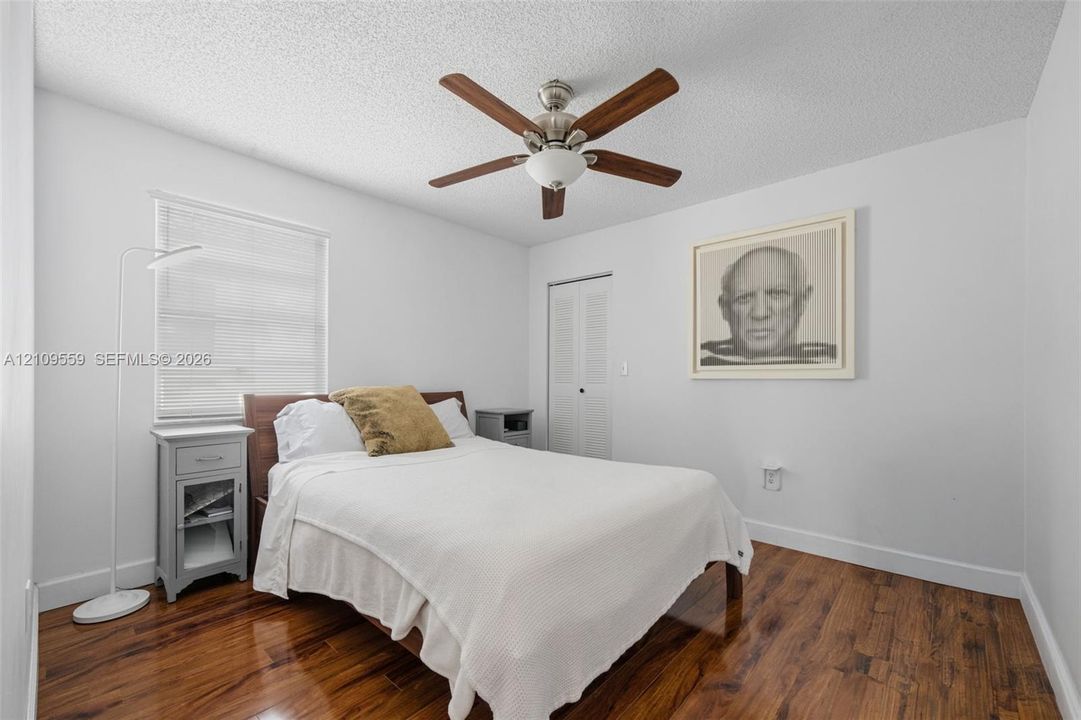 Bedroom featuring polished wood-finish flooring, a white-bladed ceiling fan with integrated light fixture, a window with horizontal blinds, and a closet with louvered double doors