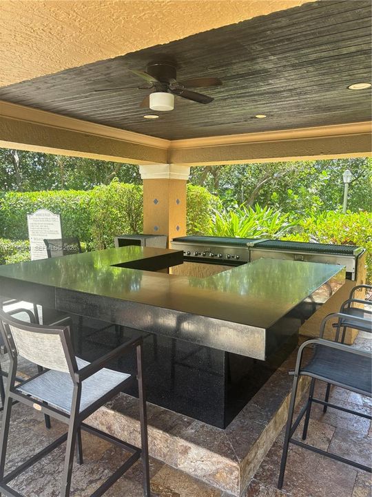 Covered outdoor kitchen featuring a stone-topped bar, stainless steel built-in grill, stone-tile flooring, and a ceiling fan