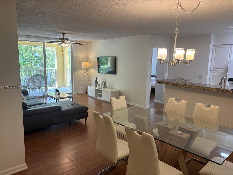 Open-concept living space with wood-finish flooring, breakfast bar with stone-surface countertop, sliding glass doors to balcony, and ceiling fan fixture