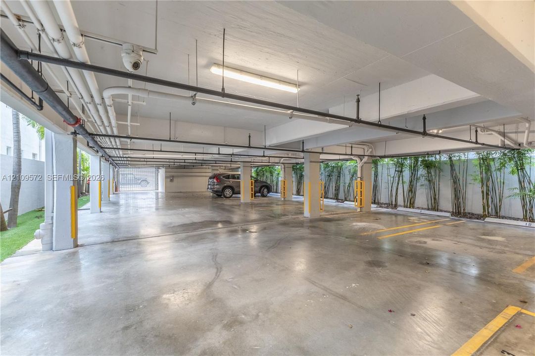 Covered parking area featuring concrete flooring, structural support columns with yellow safety bumpers, linear overhead lighting, and perimeter landscaping