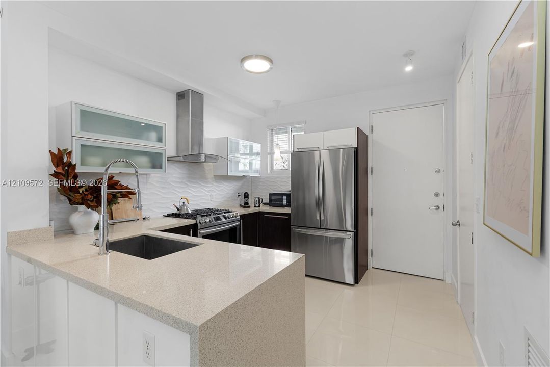 Kitchen featuring quartz countertops, stainless steel appliances, upper cabinetry with glass-front inserts, textured backsplash, and polished tile flooring