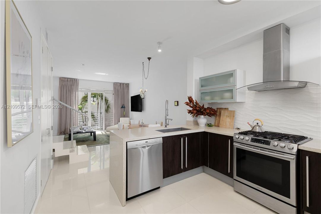 Kitchen featuring dark wood-finish cabinetry, light stone-finish countertops, stainless steel appliances, wave-patterned backsplash tile, and a wall-mounted stainless steel vent hood