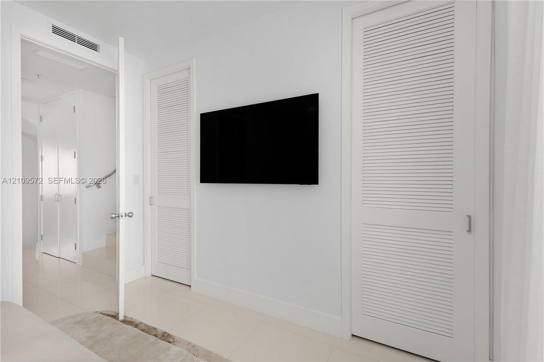 Interior space featuring white tile flooring, dual white louvered closet doors, and a wall-mounted display screen