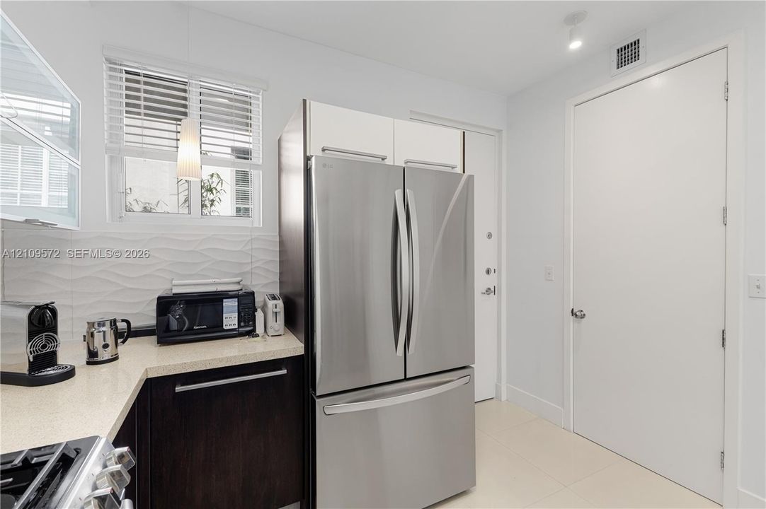 Kitchen featuring dark cabinetry, stainless steel French-door refrigerator, light-toned stone-finish countertops, textured white tile backsplash, and polished tile flooring