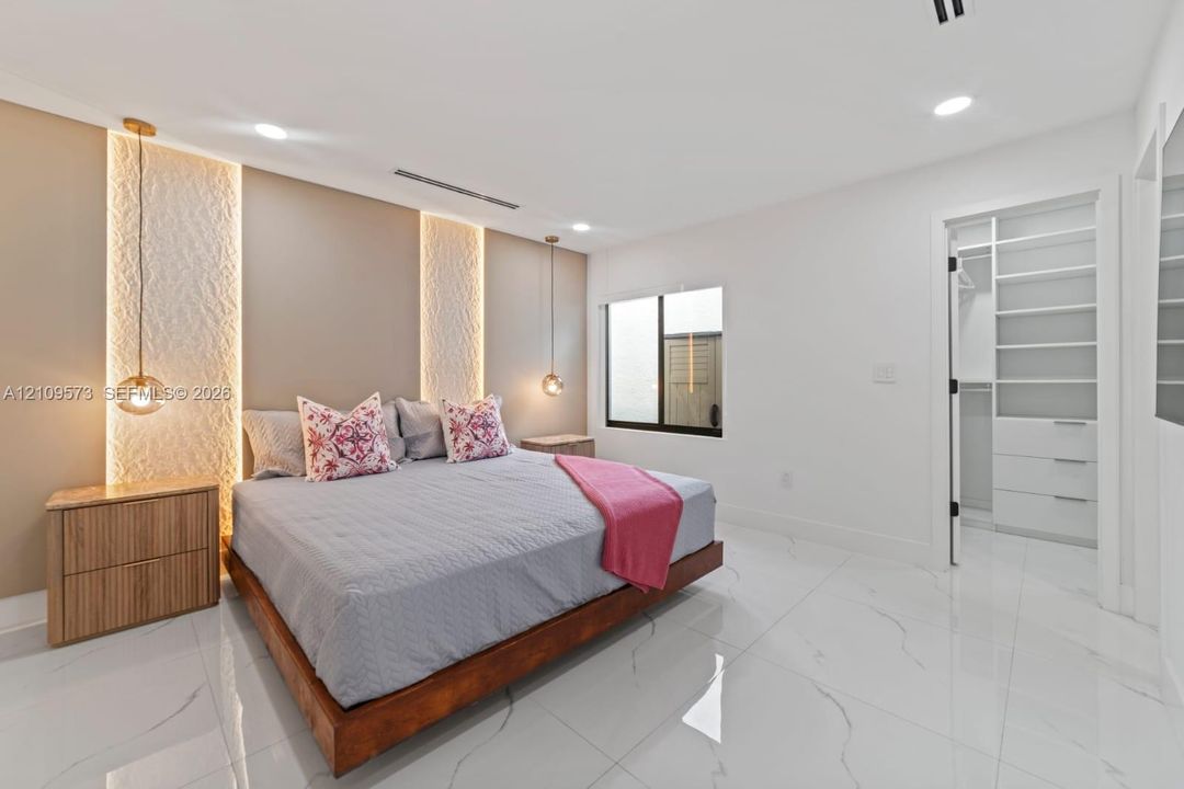 Spacious room featuring large-format tile flooring, a textured accent wall with integrated LED backlighting, recessed ceiling lighting, dual globe pendant fixtures, and a custom walk-in closet with built-in shelving