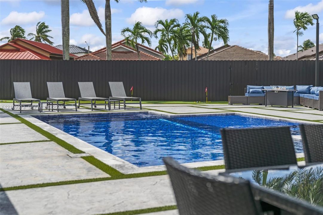 In-ground swimming pool with tile trim, surrounded by a grid-patterned concrete paver deck with synthetic turf inlays and a perimeter privacy fence