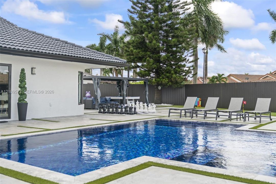 In-ground swimming pool with tile finish, expansive paver patio with turf inlays, and privacy fencing