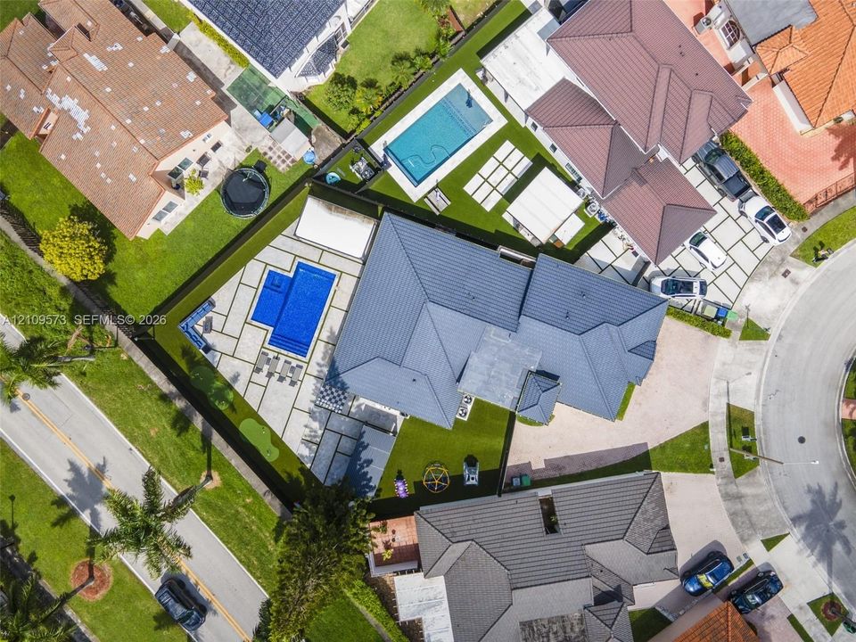 Aerial perspective of a property featuring a swimming pool, expansive paver patio, synthetic turf lawn, and a multi-faceted roof structure