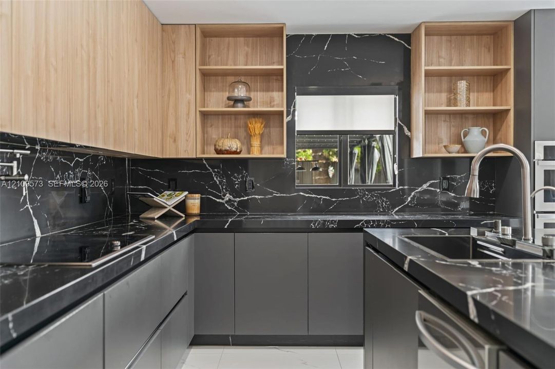 Kitchen featuring matte charcoal base cabinetry, light-toned wood-finish upper cabinets with open shelving, high-contrast stone-finish countertops, a matching full-height stone-finish backsplash, and an integrated stainless steel faucet