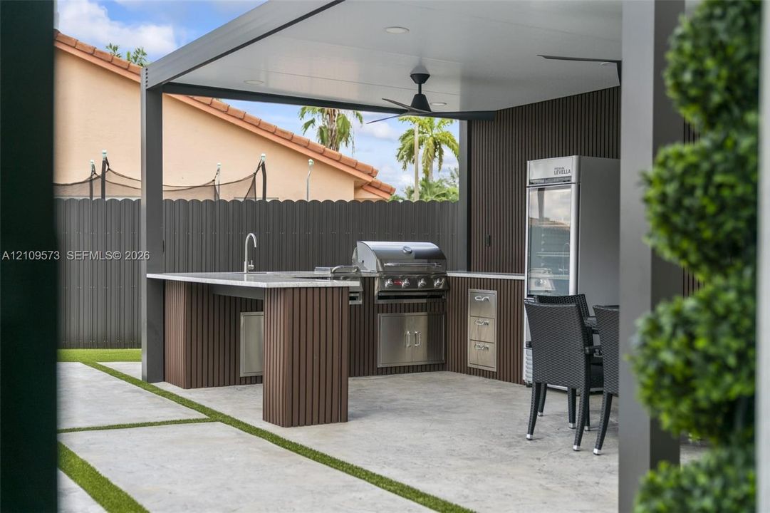 Covered outdoor kitchen featuring a stone-topped peninsula, stainless steel grill, integrated storage, and overhead ceiling fan, situated on a concrete patio with turf inlays