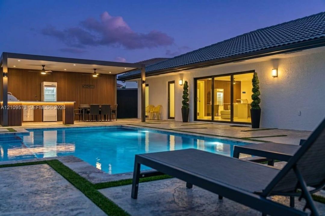 Custom swimming pool with stone-textured patio decking, integrated outdoor kitchen with wood-finish paneling, and sliding glass panel doors